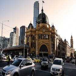 Traffic flows near the historic Flinders Street Station in Melbourne, with its distinctive yellow facade and green dome, backlit by the setting sun as Sydney lawyers navigate the urban setting.