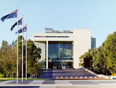 Front view of a modern building with a large glass facade, flanked by multiple flagpoles displaying windswept flags under a clear blue sky, housing prominent Sydney lawyers.