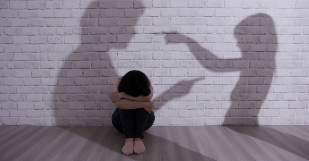 A woman sitting huddled over with a shadow of a couple behind her, suggesting a sense of fear or vulnerability