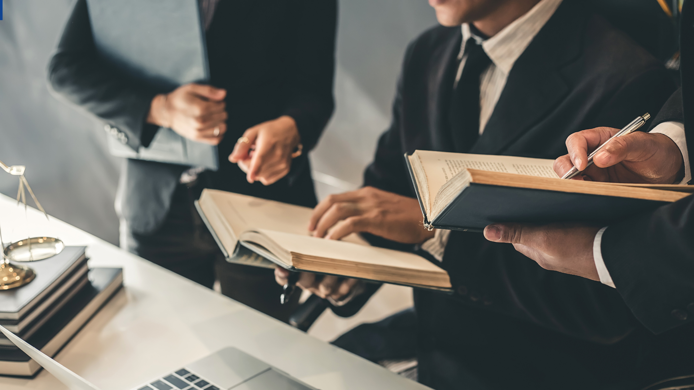 Three business professionals in a meeting, one holding an open book and taking notes, another with a laptop, in a well-lit office setting.