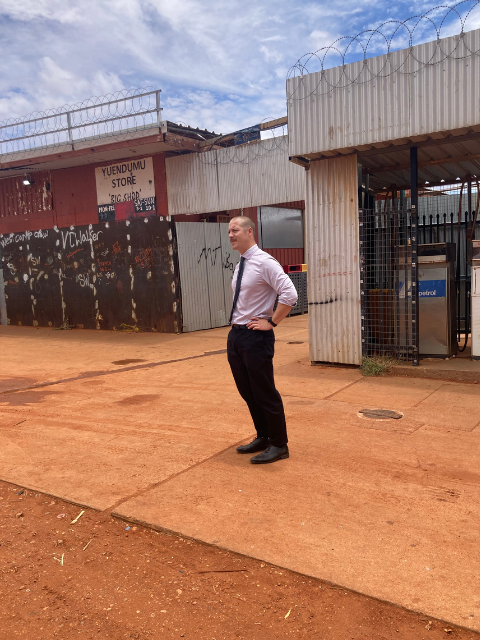 A man stands outside on a dirt pathway with his hands on his hips. He is wearing a light pink shirt, dark trousers, and dress shoes. Behind him, there's a fenced area with a secured gate and a building marked "Yuendumu Store." Razor wire tops the fence. The sky is partly cloudy.