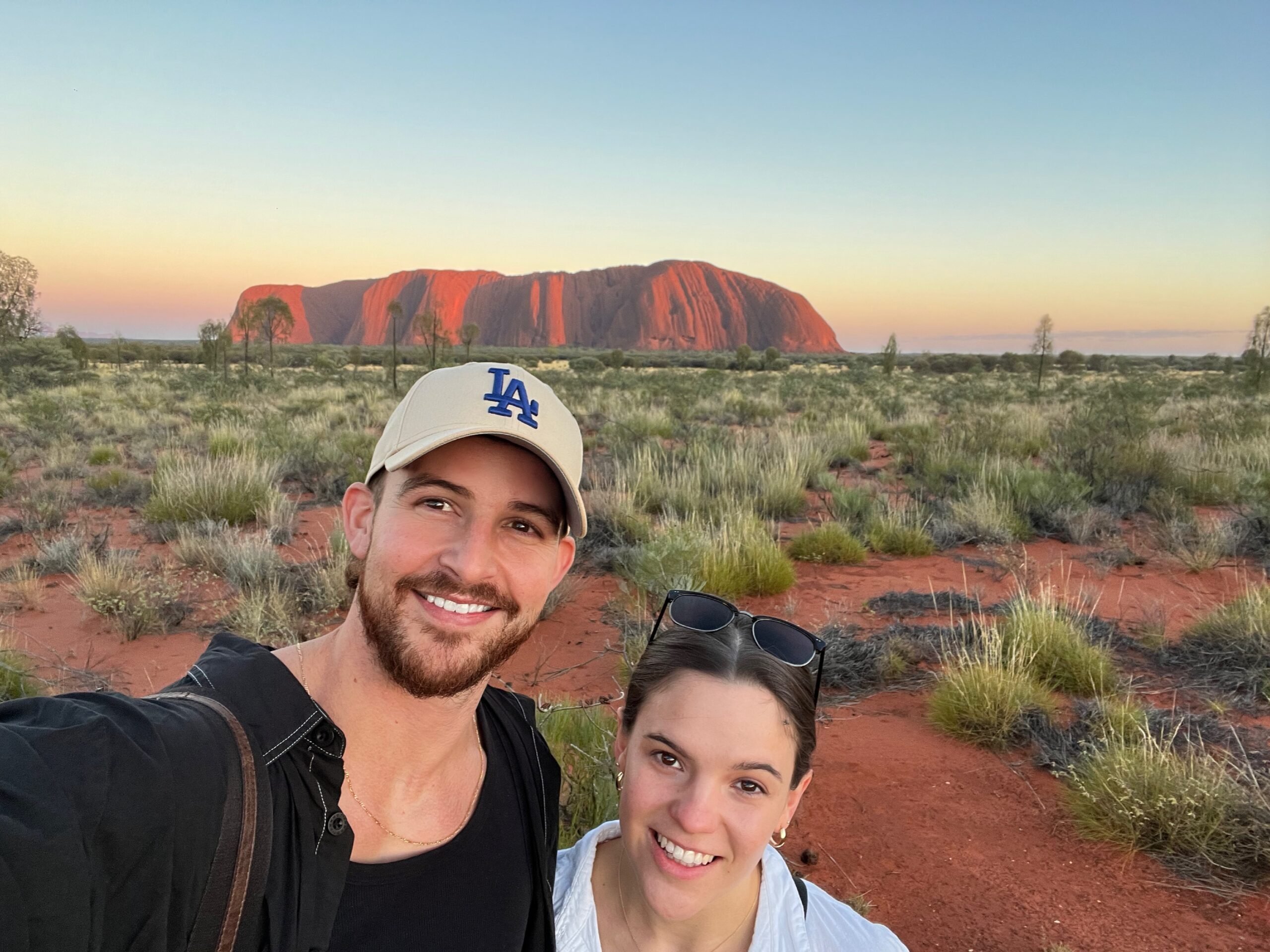 A man and woman take a selfie in a desert landscape at sunset, with a large, red rock formation in the background. The man wears a beige hat and a black shirt, the woman wears sunglasses on her head and a white shirt. Sparse vegetation surrounds them.