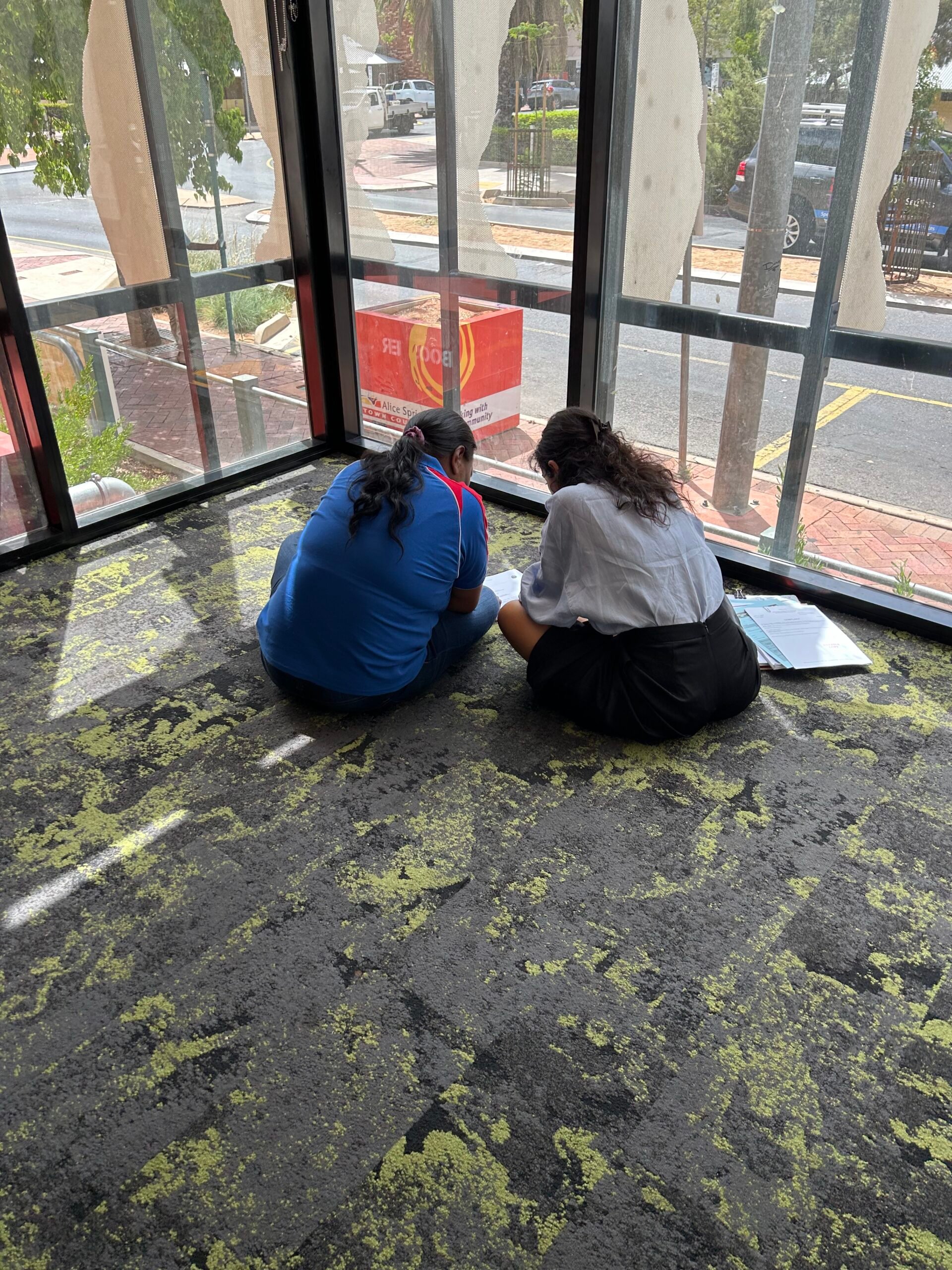 Two people sit on a carpeted floor by large glass windows. One wears a blue shirt, and the other wears a white shirt and black skirt. They are focused on a binder or document in front of them. Outside the windows, trees and parked cars are visible.