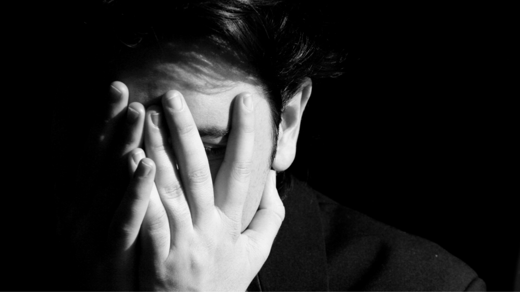 A black and white image of a person covering their face with their hands, expressing emotion or distress, set against a dark background.