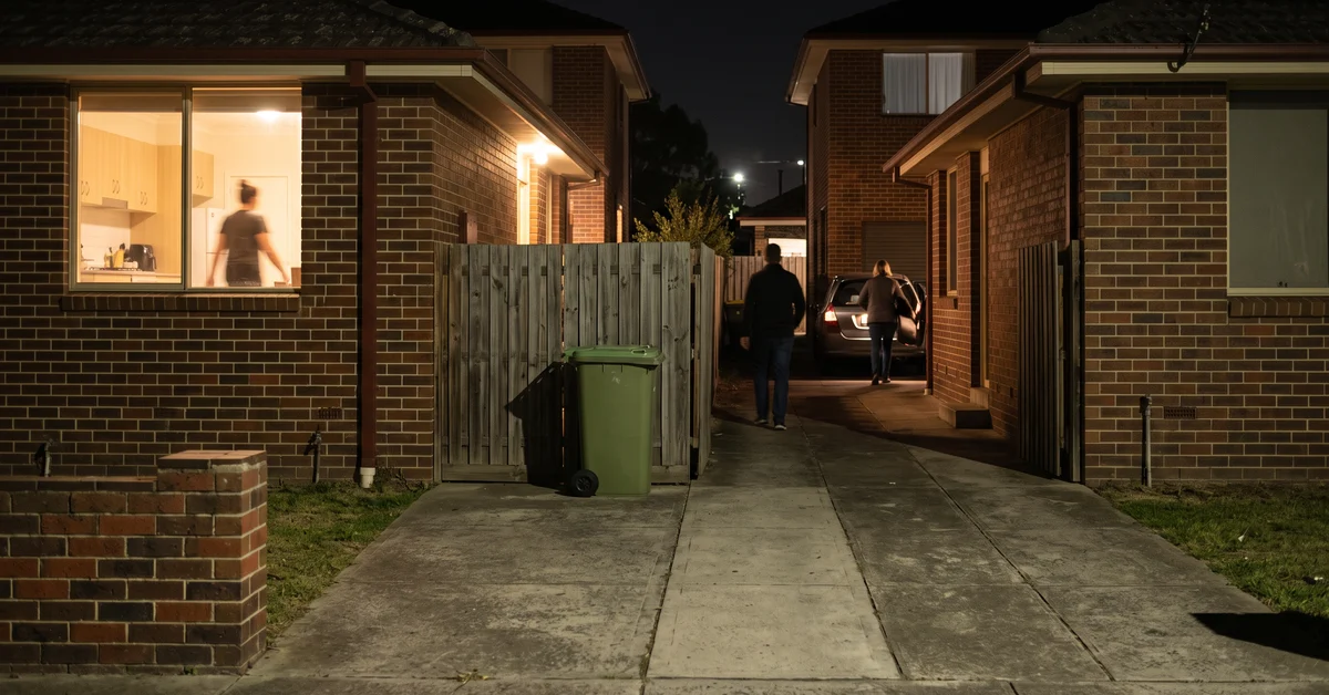 Night scene of a suburban driveway between brick houses; two people near a parked car in the distance, window showing a blurred person inside the left house.