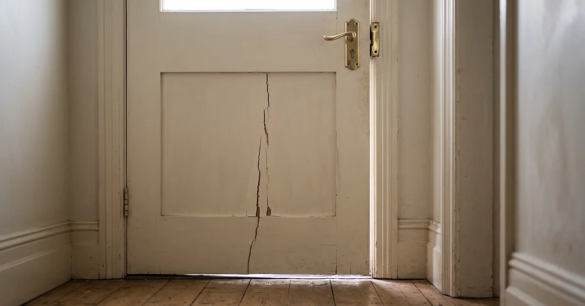 White wooden interior door with a long vertical crack and light seeping through the bottom gap in a narrow hallway floor.