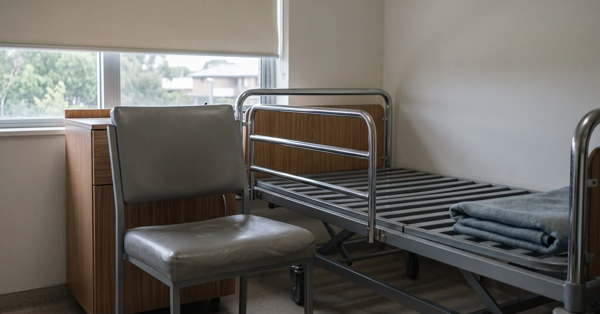 Empty hospital/dorm room with a metal-framed bed, gray chair, and a wooden dresser by a window; a folded blue blanket sits on the bed frame.