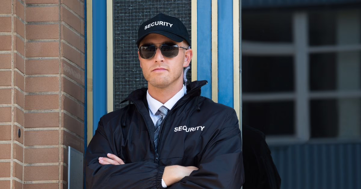 Suing Security Guard For Assault, Battery & False Imprisonment In NSW_ Birthday Brawl (2) | O'BRIEN CRIMINAL & CIVIL SOLICITORS A security man stands with crossed arms in front of a brick wall and blue door wearing a black baseball cap and uniform