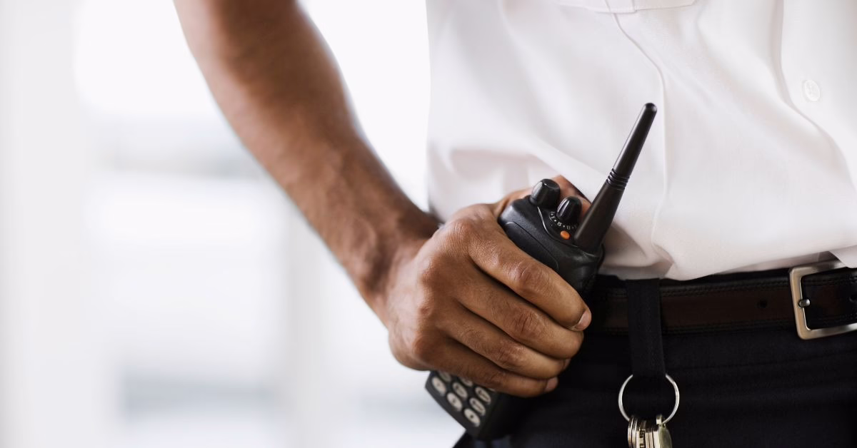 A security guard's hand on his hip holding a walkie talkie wearing a white shirt and black trousers