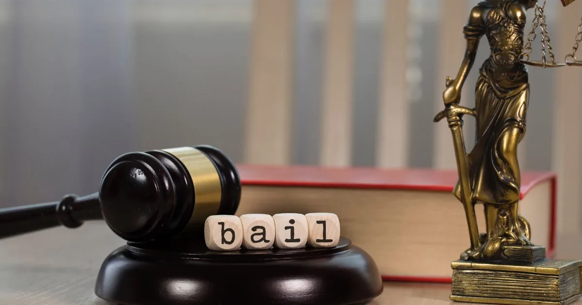 Wooden blocks spelling "bail" beside a judge's gavel, a red law book and a brass Lady Justice statue holding scales. 