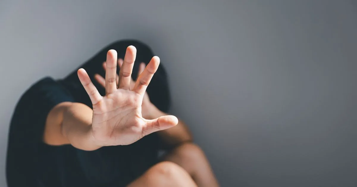 Person sitting against a gray wall raises a hand in a stop/shielding gesture, face blurred behind the hand!