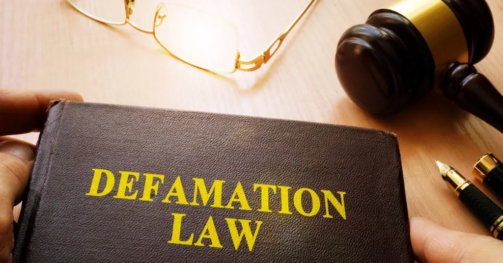 A dark leather book titled 'Defamation Law' in yellow letters on a wooden desk, with a gavel, fountain pens, and glasses nearby.