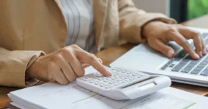 Person using a calculator while typing on a laptop, with financial papers on a desk—conveying budgeting or accounting work.